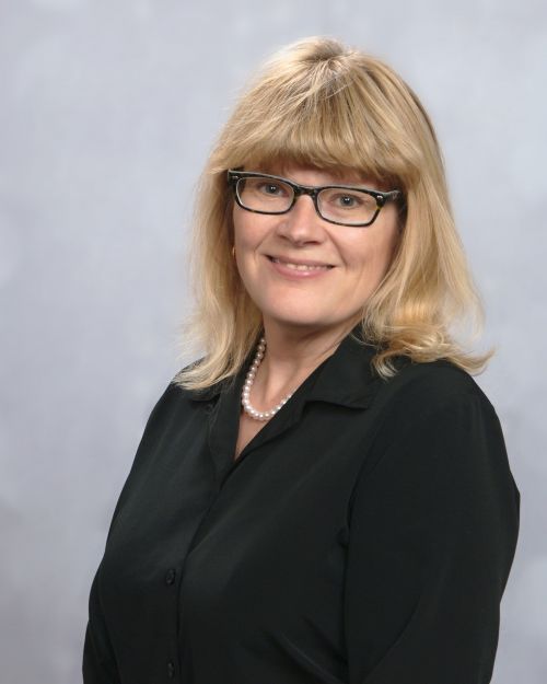 Image of Rebecca Lehto has blonde hair and glasses smiles at camera with portrait backdrop.